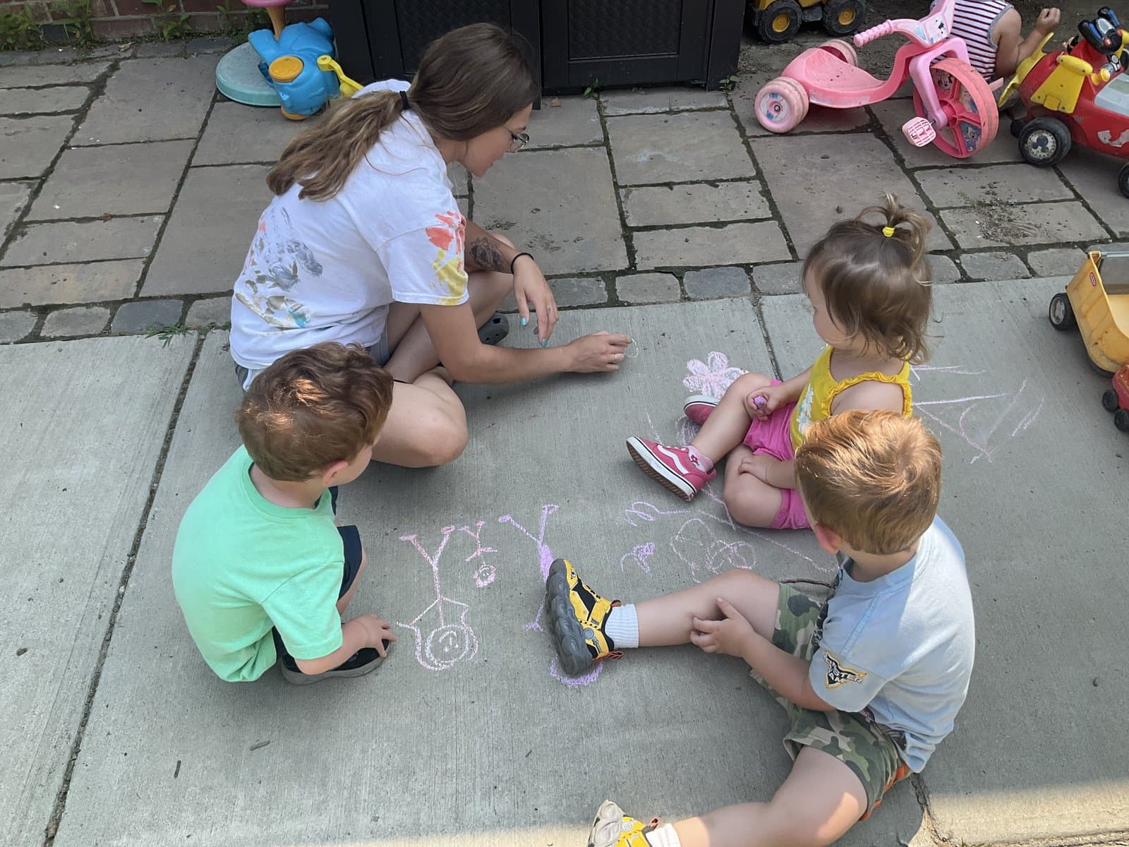 Little Acorns teacher drawing chalk art on the sidewalk with three children outside